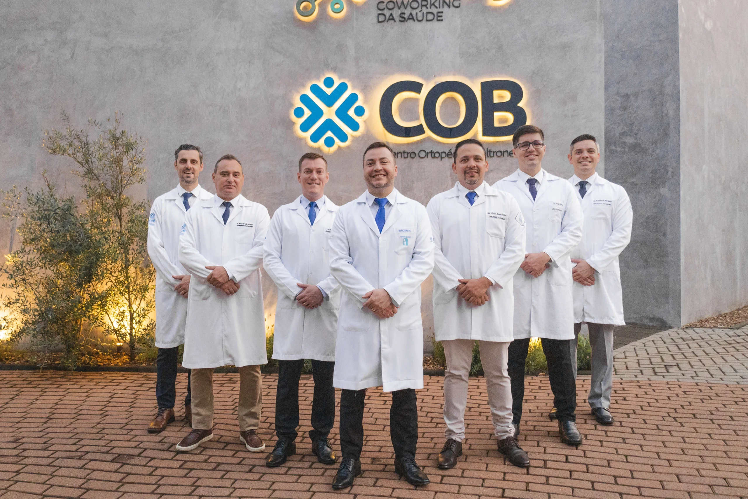 Grupo de profissionais médicos, todos vestidos com jalecos brancos e gravatas azuis, posando sorridentes em frente ao logotipo do "COB - Centro Ortopédico Beltrame". O fundo exibe uma parede cinza com elementos de paisagismo iluminados, criando um ambiente moderno e acolhedor.